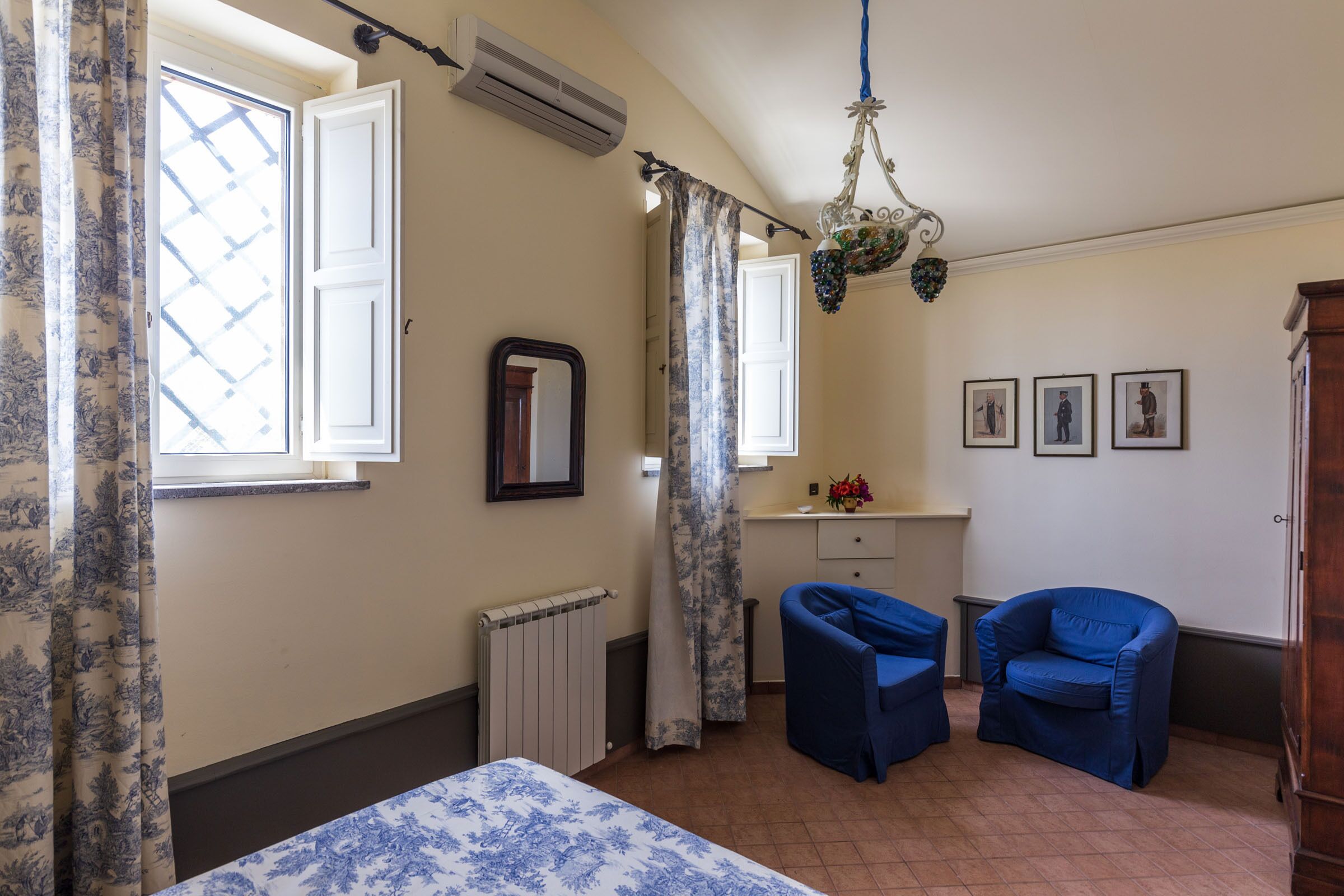 A bedroom with a bed, two blue chairs, a radiator, a mirror, and a window with curtains.