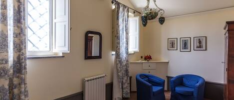 A bedroom with a bed, two blue chairs, a radiator, a mirror, and a window with curtains.