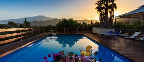 Un espace aménagé au bord de la piscine, avec une terrasse en bois, des chaises longues et une table où sont disposées des boissons et des fleurs.