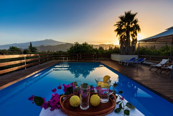 A poolside setting with a wooden deck, lounge chairs, and a table with drinks and flowers.