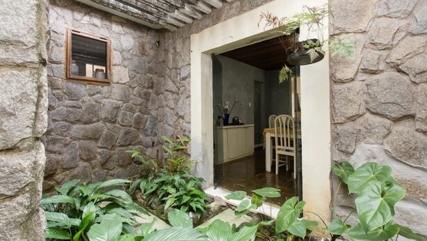 Een gebouw met stenen muren, een klein raam en een deuropening naar een kamer met een stoel en een tafel, met weelderige groene planten op de voorgrond.