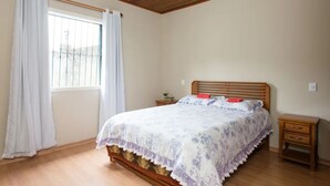 Een slaapkamer met een houten plafond, een bed met een bloemenprintdeken, een nachtkastje en een raam met gordijnen.