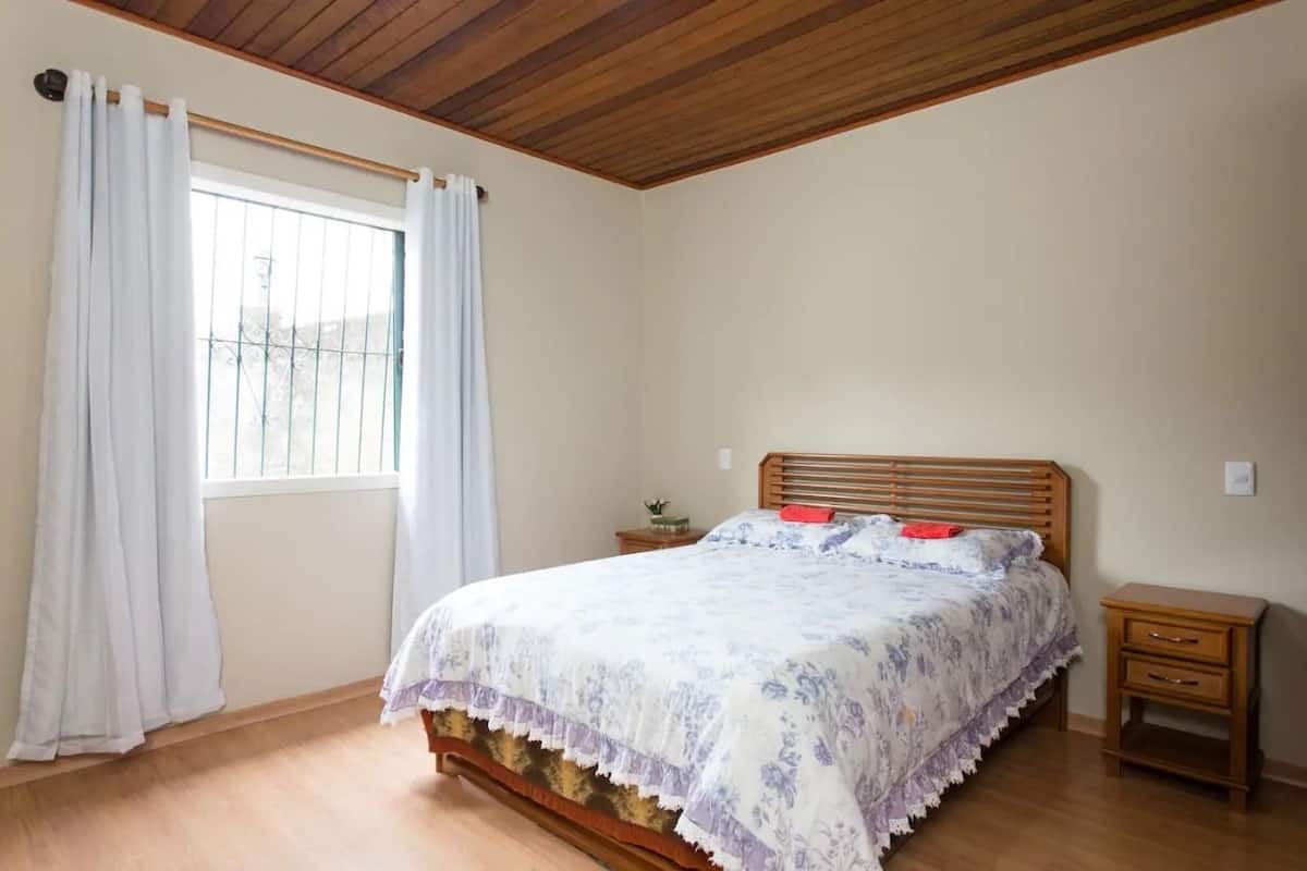Ein Schlafzimmer mit Holzboden, einem Bett mit floralem Überwurf, einem Nachttisch und einem Fenster mit Vorhängen.