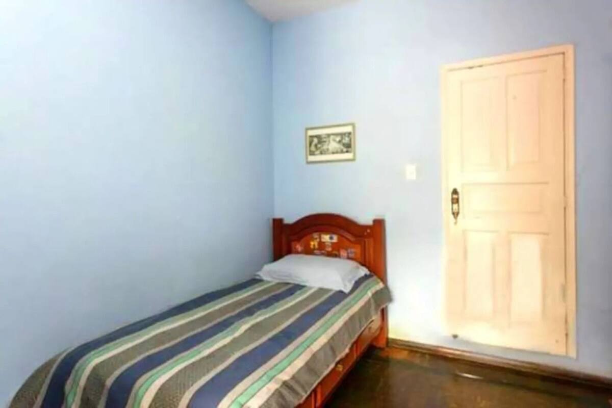 Ein kleines Zimmer mit einem Einzelbett, einem Holz-Kopfteil, einem gerahmten Bild an der Wand und einer geschlossenen Tür.