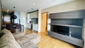 A modern living room with a large sectional sofa, a flat-screen TV mounted on a wooden wall, and a kitchen in the background. - Apartment in front of Fira Gran Via (Hospitalet de Llobregat - Barcelona)