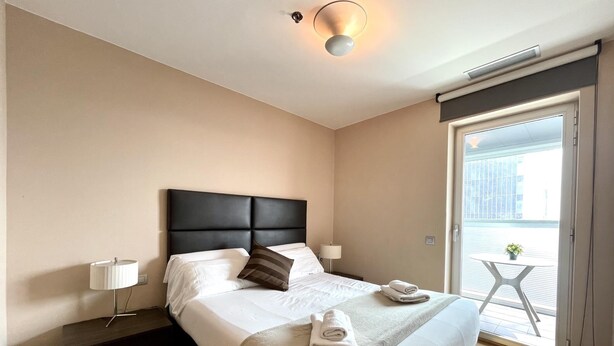 A modern hotel room with a large bed, a bedside table, a lamp, and a view of the outside balcony. - Apartment in front of Fira Gran Via (Hospitalet de Llobregat - Barcelona)