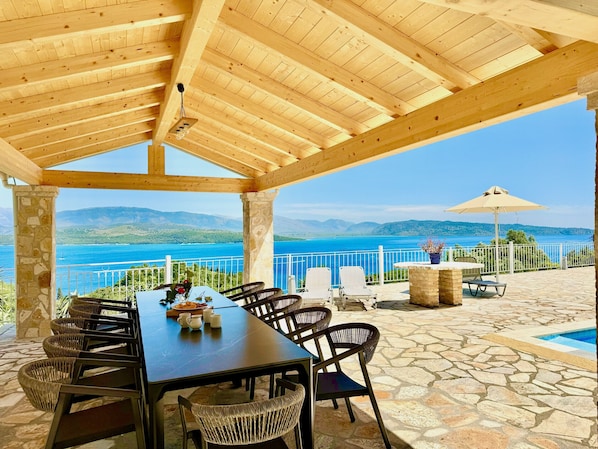 A covered outdoor dining area with a table and chairs, overlooking a body of water and mountains.