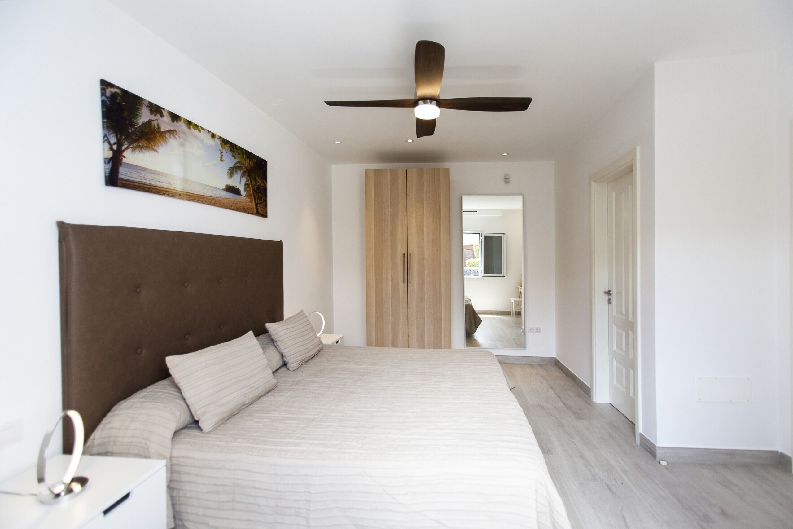 Une chambre avec un grand lit, un ventilateur de plafond, une armoire en bois et un tableau accroché au mur.