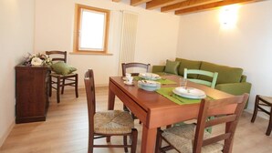 Zona pranzo con tavolo in legno apparecchiato per quattro, circondato da sedie in legno con sedute intrecciate. Contro il muro si trova un divano verde, e si vede una finestra con persiane.