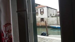 Une vue à travers une fenêtre sur un canal où un bateau est amarré, un bâtiment avec un toit recouvert de tuiles rouges et un mur blanc avec des fenêtres.