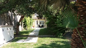 Un chemin menant à une maison avec un jardin, un palmier et un petit abri blanc servant d'abri de jardin.