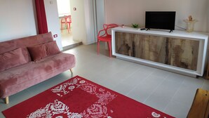 Un salon avec un canapé rose, un tapis rouge à motifs, une console blanche sur laquelle se trouve un téléviseur, et un fauteuil rouge.