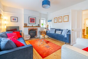 A living room with a sofa, armchair, fireplace, and a door leading to another room. - Sybil Cottage (4), Holme-next-the-Sea, Norfolk (Holme-next-the-Sea)