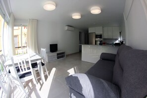 A modern living room with a grey sofa, a dining table with white chairs, a TV on a wooden stand, and a kitchen in the background.
