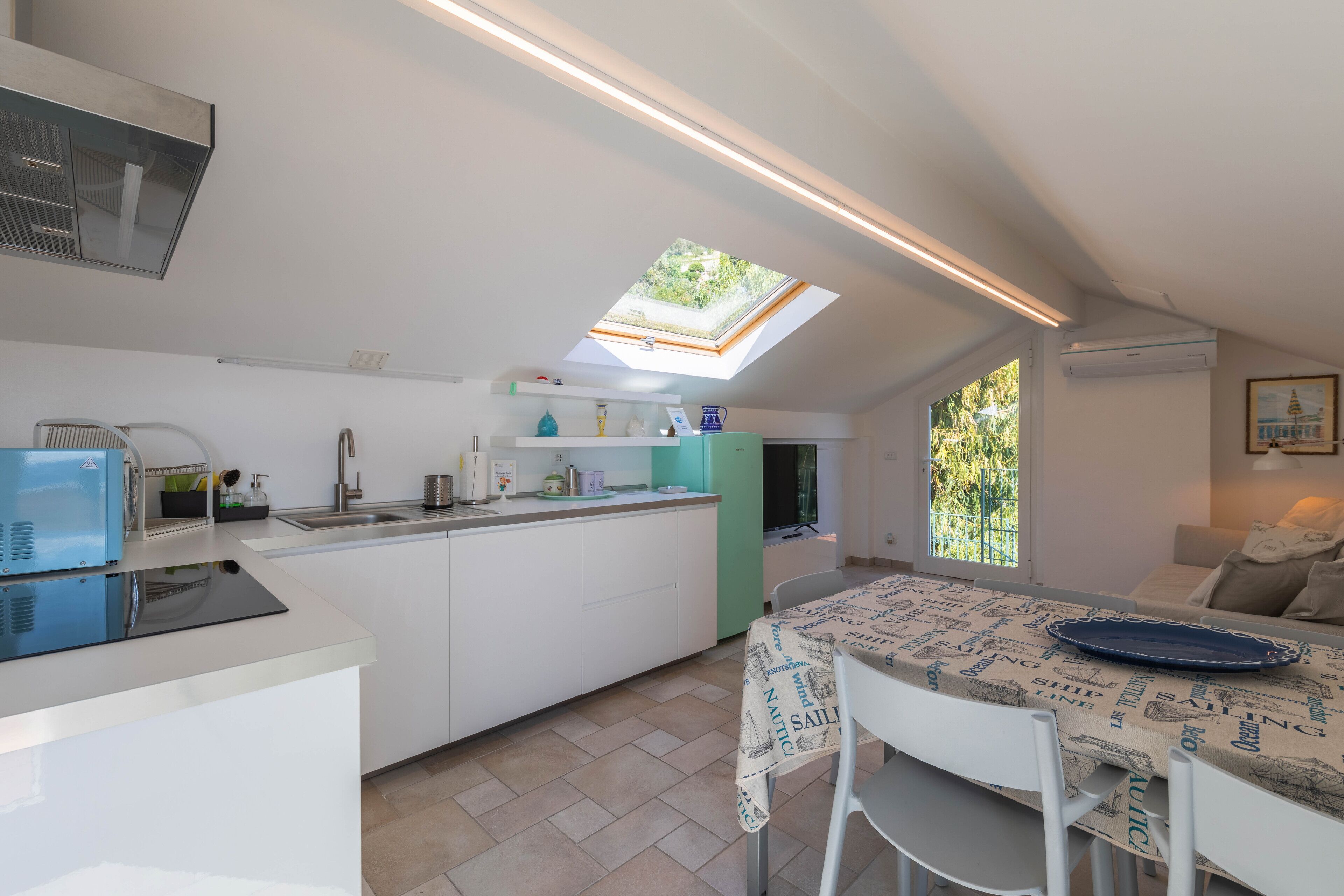 A modern kitchen with a skylight, white cabinetry, and a dining area with a table covered in a nautical-themed tablecloth.