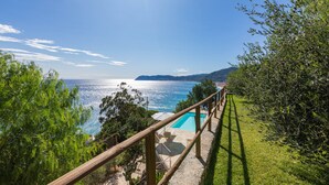Zona de piscina con terraza de madera, rodeada de abundante vegetación y vistas panorámicas al mar y montañas distantes.