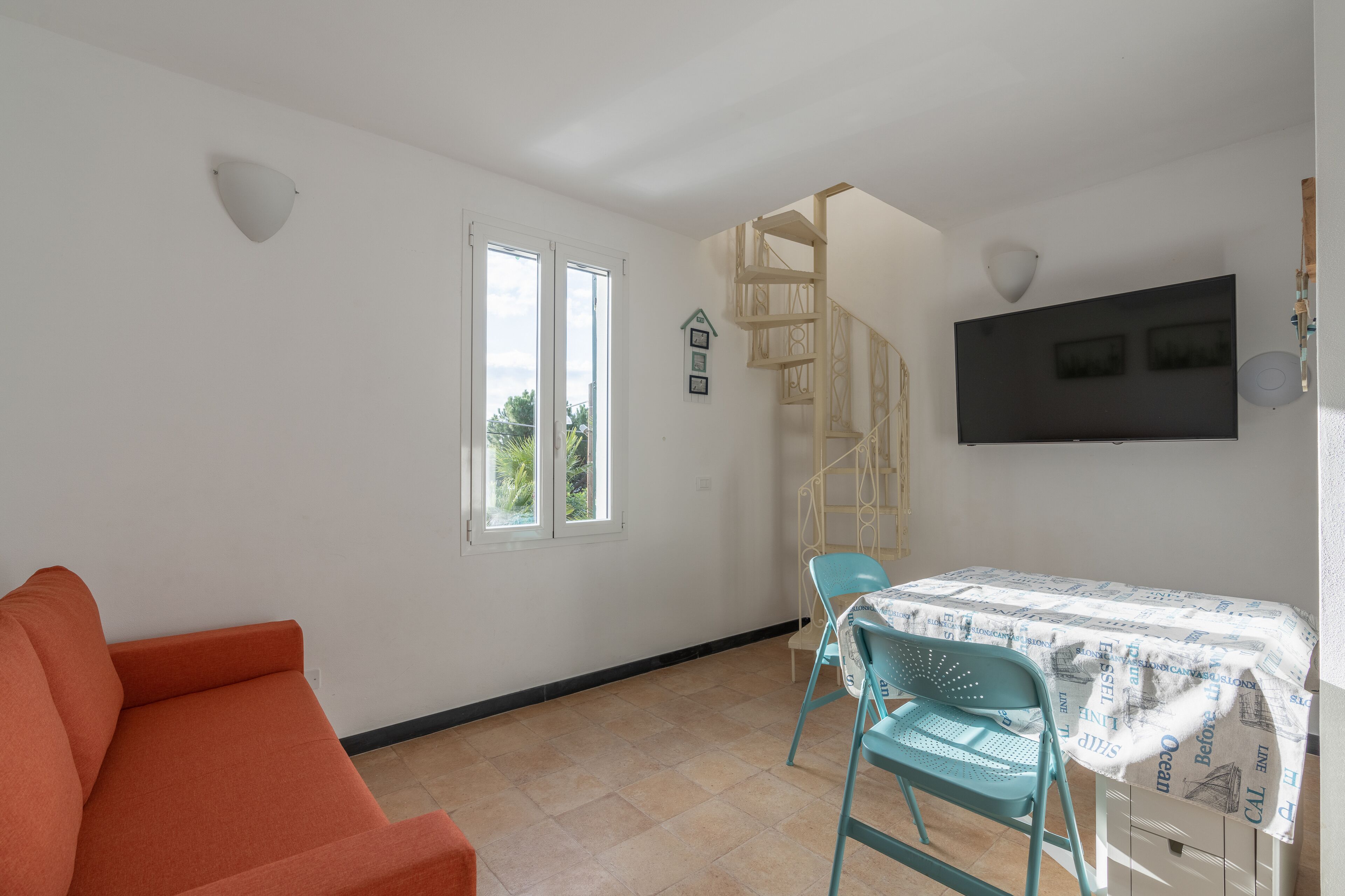 A room with a sofa, a table with a tablecloth, a chair, a staircase, and a TV mounted on the wall.
