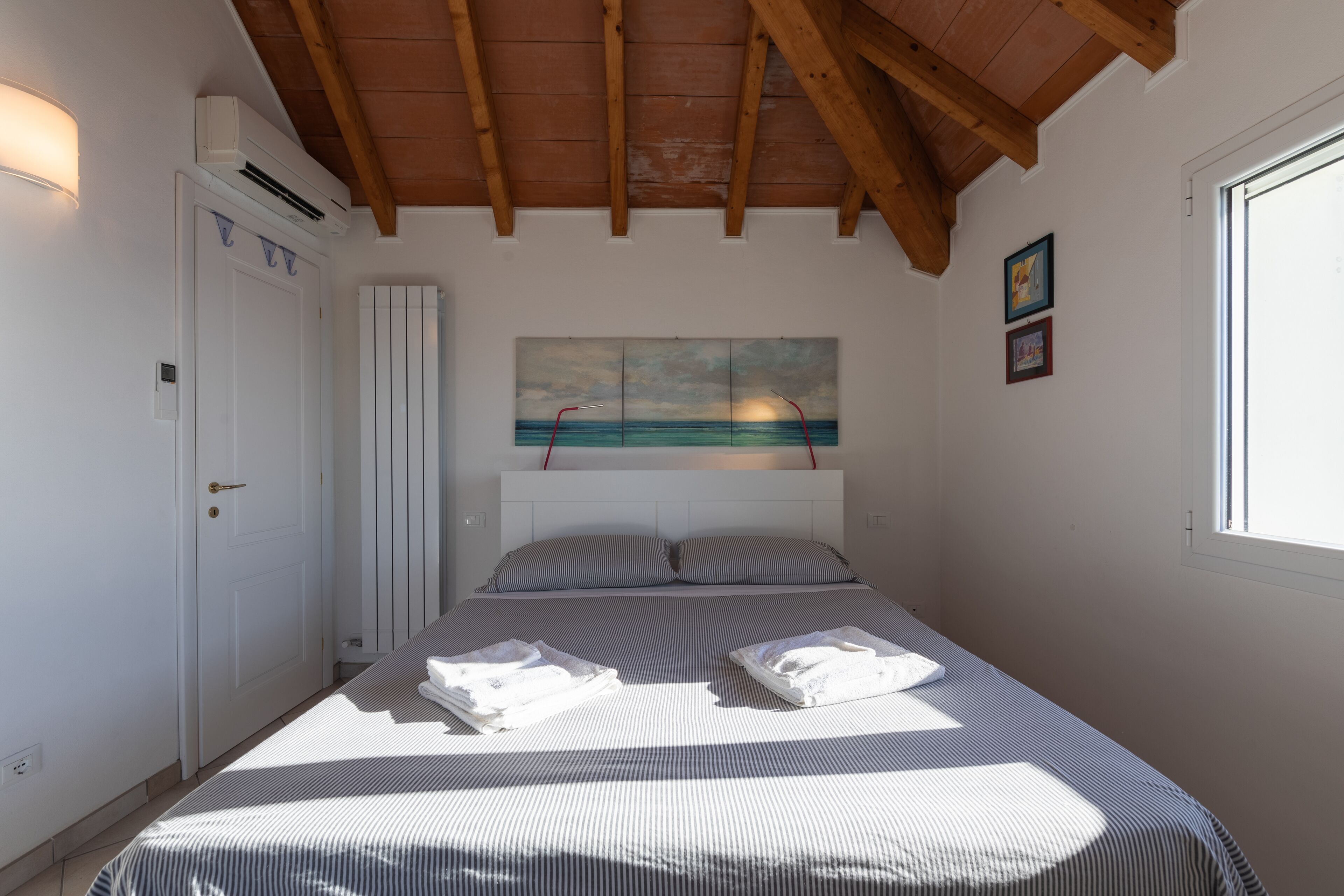 A bedroom with a bed, a white door, an air conditioning unit, a painting, and a window.