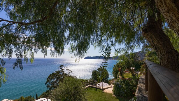 Vista costera con terraza de madera, zona verde y una perspectiva despejada del mar y la costa distante.
