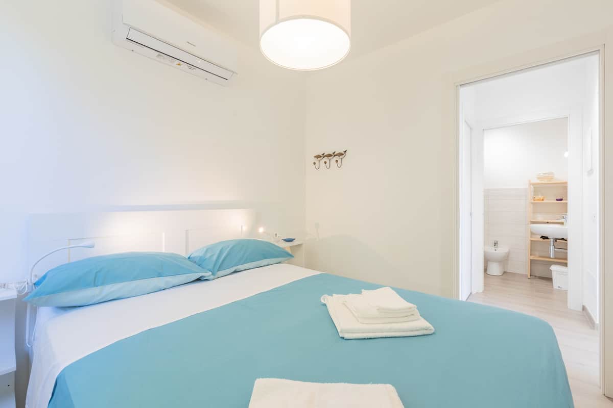 A neatly made bed with blue bedding, a bedside table with a lamp, and a wall-mounted air conditioner.