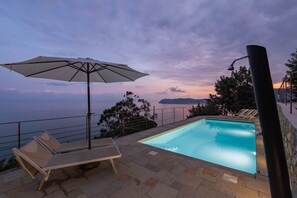 Zona piscina con una panchina, un ombrellone e vista sul mare al crepuscolo.