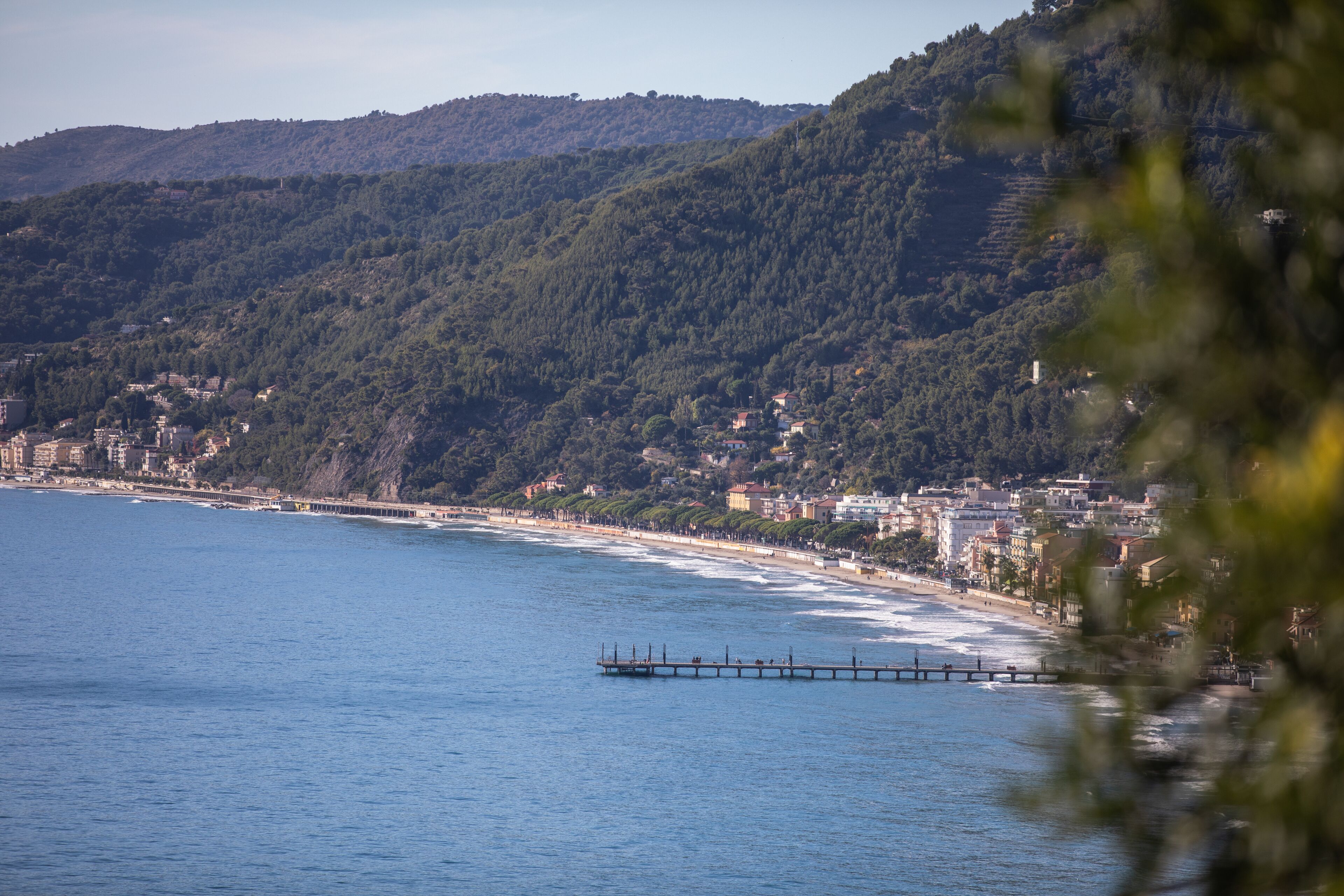 A coastal town with a pier extending into the sea, surrounded by hills and buildings.