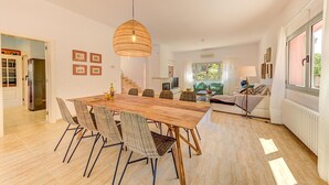 A spacious living room with a large wooden dining table, wicker chairs, a hanging light fixture, and a view of the outdoors through large windows. - Maria Bonaire for eigth people with pool (Alcúdia)
