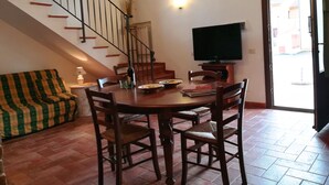 Un comedor con una mesa de madera, sillas y un televisor. Se observa una escalera con barandilla y una ventana. Un sofá es visible en una esquina.