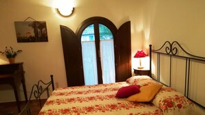 Un dormitorio con una cama, una mesita de noche, una lámpara, un cuadro en la pared y una ventana con puerta abierta.
