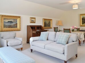 A living room with a light-colored sofa, a matching armchair, and a bench. There are two wooden dressers, a table with a floral tablecloth, and a lamp.