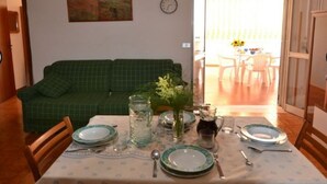 Ein Essbereich mit Tisch für vier Personen, einem grün karierten Sofa, einer Wanduhr und Blick ins Freie durch eine Glastür.