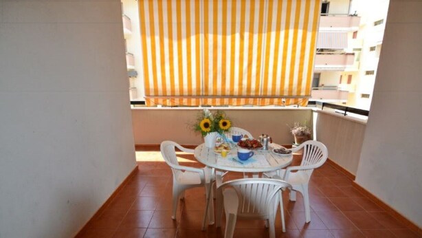 Ein Balkon mit rundem Tisch und vier Stühlen, einem Streifenmarkisenvorzelt und Blick auf ein Gebäude im Hintergrund.
