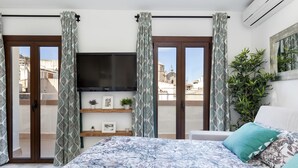 Ein Schlafzimmer mit einem großen Bett, einem Flachbildfernseher, einem kleinen Regal mit Pflanzen und Bildern sowie einem Balkon mit Blick auf Gebäude.