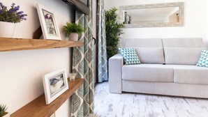 Ein modernes Wohnzimmer mit einem grauen Sofa, Holzregalen mit gerahmten Bildern, einem großen Spiegel und einem gemusterten Vorhang.