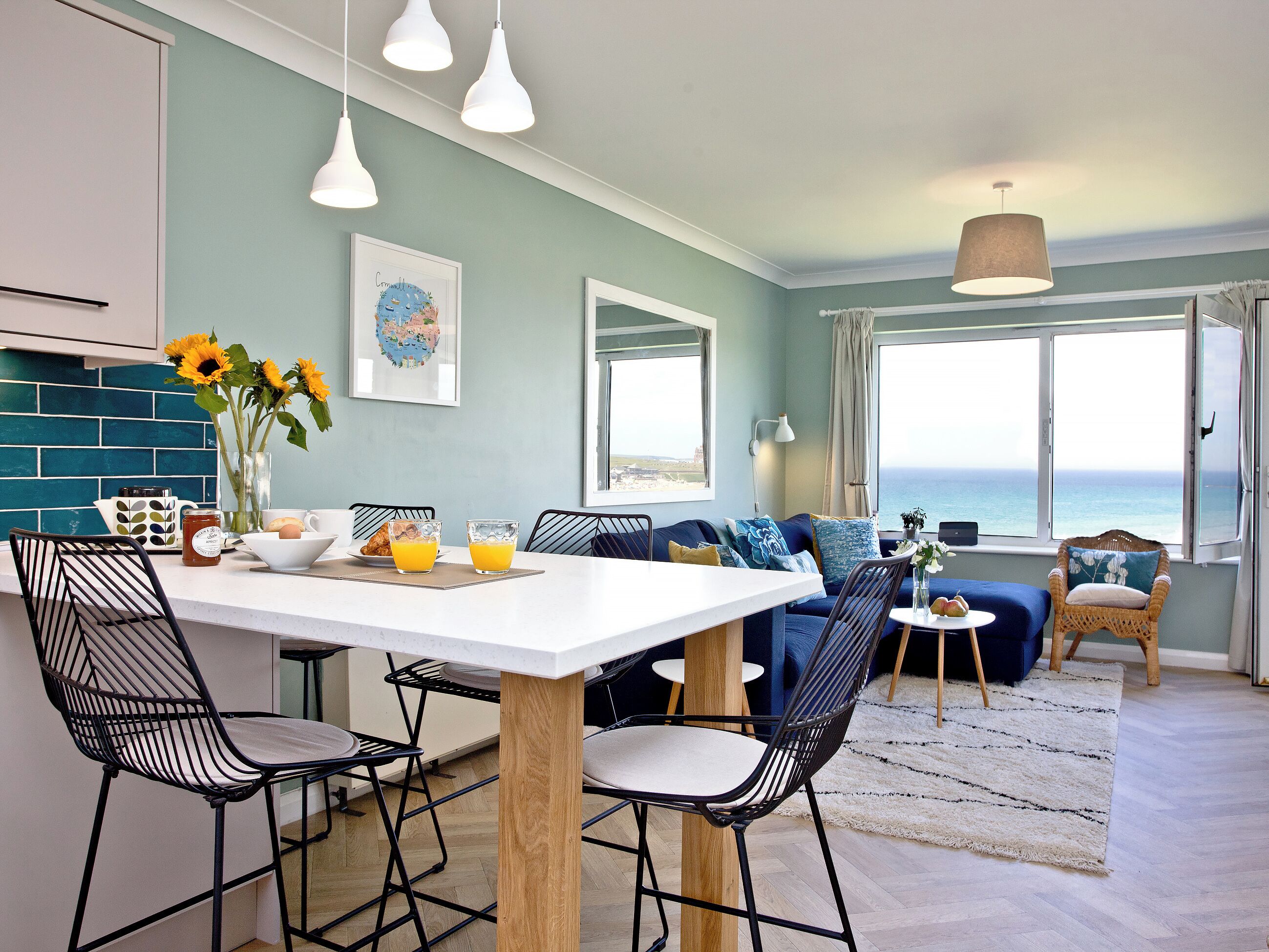 A modern kitchen with a white countertop, black chairs, and a dining table. There is a large window with a view of the ocean.