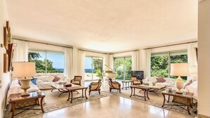 Amplia sala de estar con televisor grande, varios rincones de descanso y ventanas amplias con vistas al exterior.
