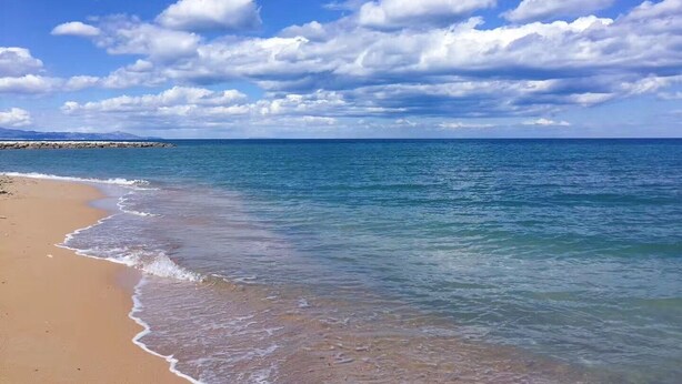 A beach with clear blue water, a sandy shore, and a rocky breakwater in the distance. - Denia beach, ideal for families (Dénia)