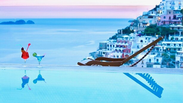 Vista della piscina con una sedia a sdraio e una persona che sorseggia una bevanda, affacciata su una cittadina costiera dai colori vivaci.