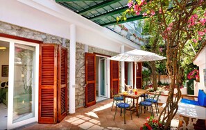 An outdoor patio with a table and chairs, a glass door, and a tree with pink flowers. - IL GIARDINO DI POSITANO (Positano)