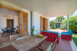 Eine überdachte Terrasse mit einem Holztisch und Stühlen, einem roten Sofa und einem Esstisch, der mit einer Blumen- und Obstschale dekoriert ist.