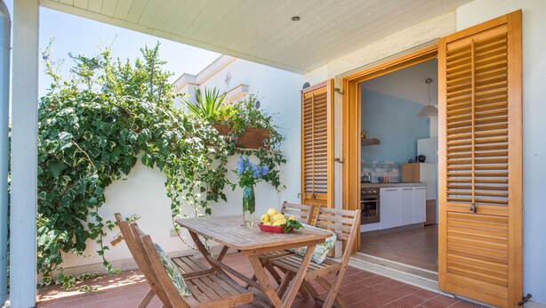 Um pátio externo com mesa e cadeiras de madeira, um cesto de limões e vista para a cozinha através de portas de madeira abertas.