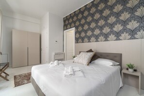 A neatly made bed with white linens and towels, a bedside table with a plant, and a patterned wallpaper on the wall. - Room 1 - Grifoni Boutique Hotel (Venice)