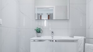 Un baño moderno con baldosas de mármol blanco, un tocador blanco con espejo, una planta en maceta y un cubo de madera.