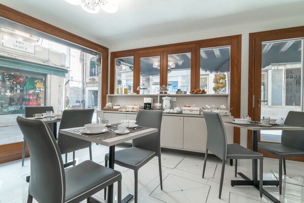 Una cafetería con una decoración moderna, que presenta una barra central con una máquina de café, una variedad de alimentos para el desayuno a la vista y varias mesas y sillas para sentarse.