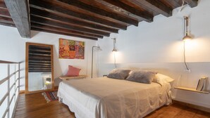 A bedroom with a wooden ceiling, a bed with a white bedspread, two bedside lamps, a small bedside table, a colorful painting on the wall, and a window with blinds. - Cozy and modern studio apartment by Mmega (Florence)