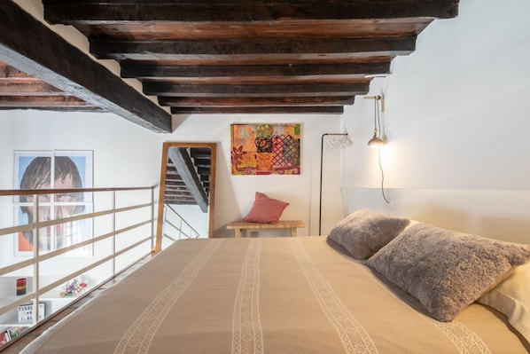 A bedroom with a bed, a wooden bench, a colorful painting, and a staircase.