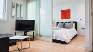 Ein modernes Hotelzimmer mit einem großen Flachbildfernseher, einem Bett mit weißer Bettwäsche, einem kleinen Tisch mit Marmorplatte und einer Schiebetür zu einem Balkon.