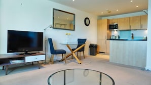 Ein modernes Wohnzimmer mit Flachbildfernseher, Glastisch und Essbereich mit blauen Stühlen.