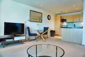 A modern living room with a flat-screen TV, a glass coffee table, and a dining area with blue chairs.