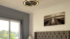A hotel room with a large bed, a nightstand, a lamp, a ceiling fan, a window with curtains, and a wall-mounted picture. - Barri Vacation Home (Kingston)